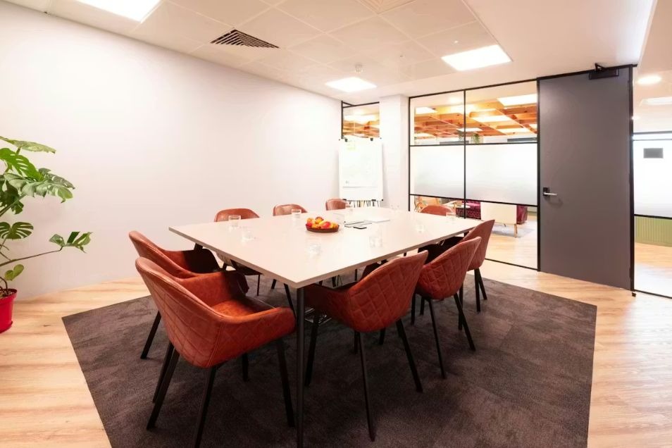 Meeting room with a light square table, rust-coloured quilted chairs, a dark area rug, and wood-look flooring.