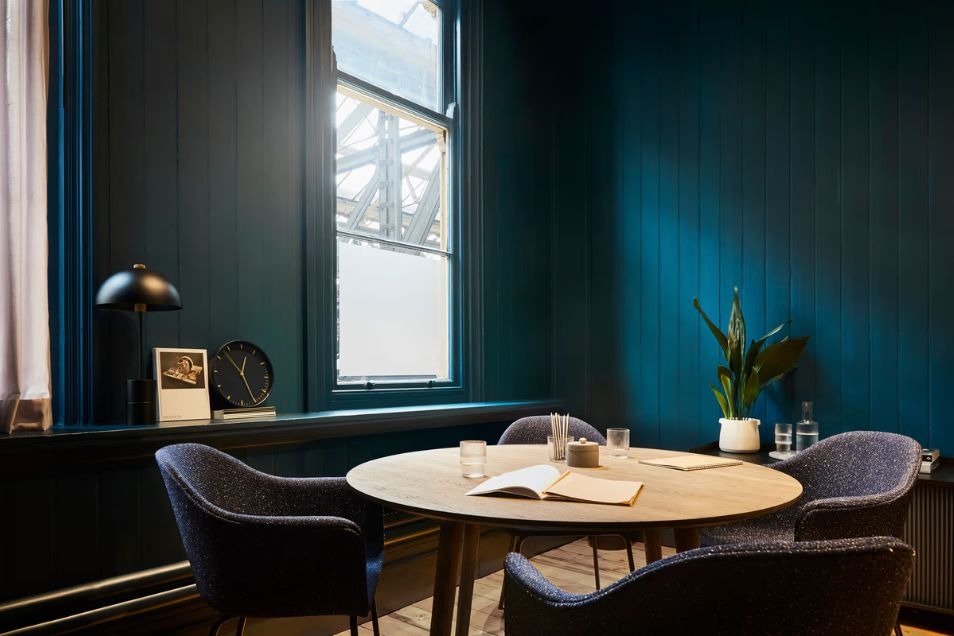 Intimate meeting room with a round wooden table, navy upholstered chairs, and deep blue wood-panelled walls by a window.