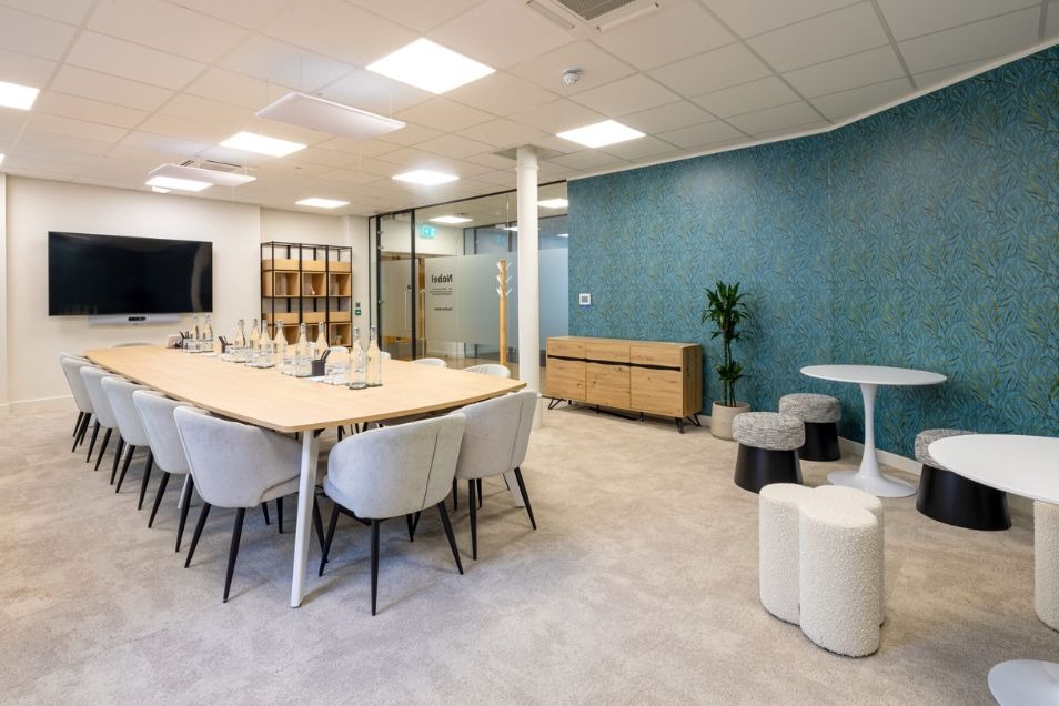 Spacious meeting room with a long light-wood table, grey tub chairs, and a teal patterned feature wall with casual cafe seating.