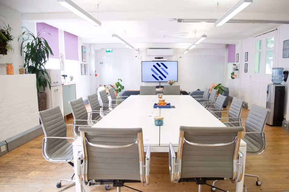 Bright meeting room with a large white boardroom table, grey ribbed leather chairs, wood floors, and lavender window blinds.