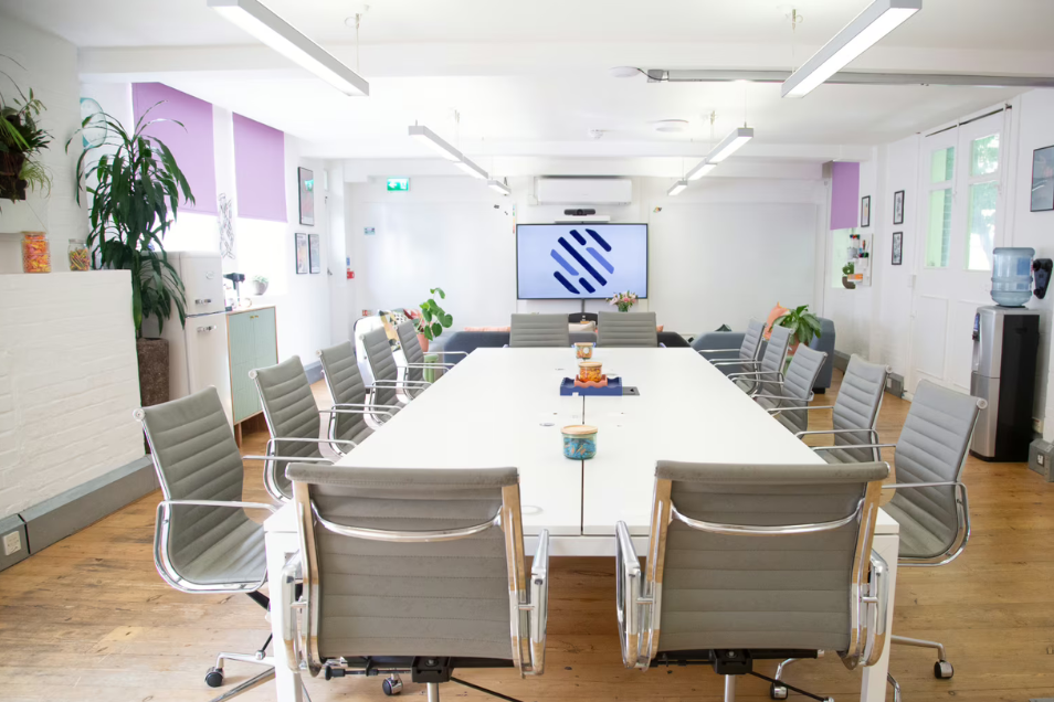 Bright meeting room with a large white boardroom table, grey ribbed leather chairs, wood floors, and lavender window blinds.
