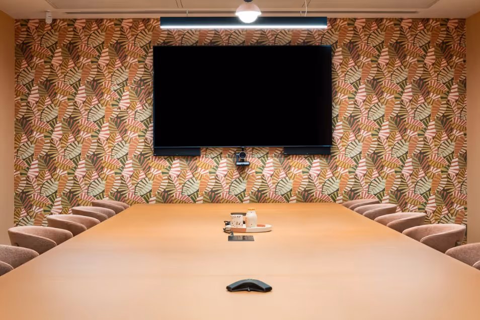 Warmly-lit boardroom with a long tan table, neutral chairs, and a vibrant botanical-patterned feature wall with a large mounted screen.