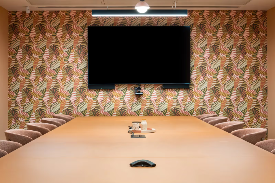 Warmly-lit boardroom with a long tan table, neutral chairs, and a vibrant botanical-patterned feature wall with a large mounted screen.