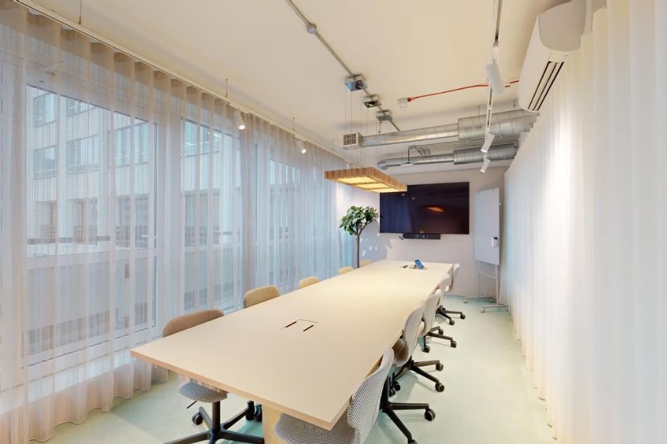 Bright, modern boardroom featuring a long blonde wood table, grey swivel chairs, industrial ceiling details, and floor-to-ceiling sheer white curtains.