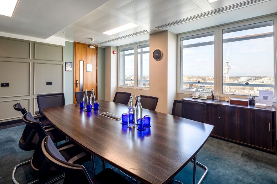 Professional meeting room with a dark wood table, blue glassware, grey panelled walls, and windows offering an elevated city view.
