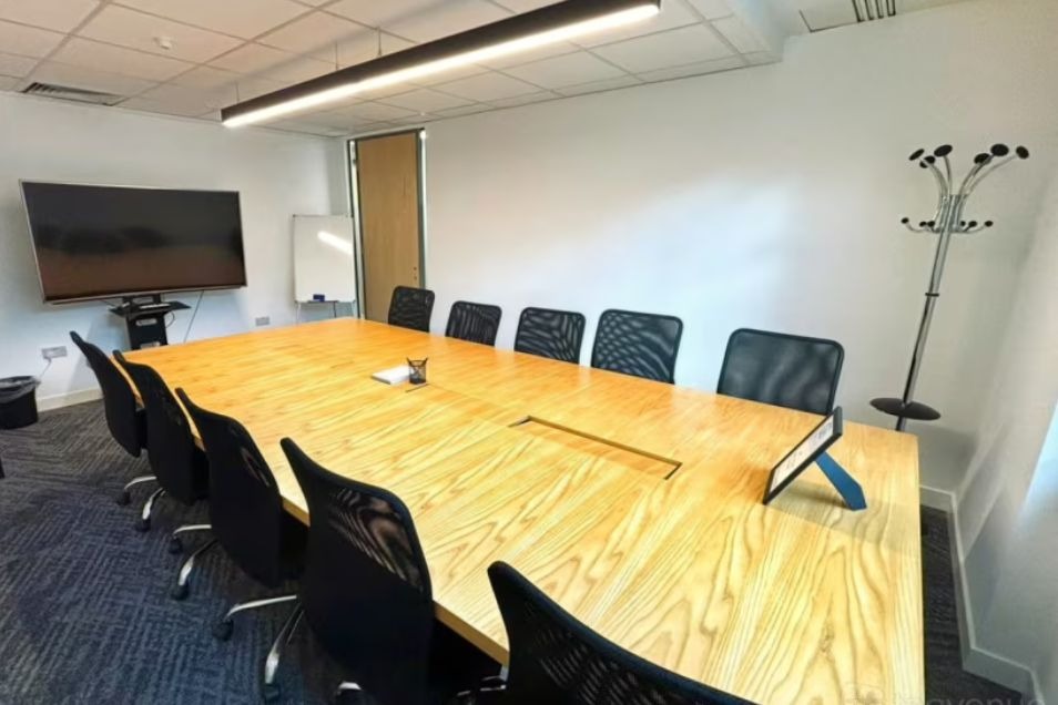 Boardroom with a light wood table, black mesh chairs, a large monitor on a stand, and a metal coat rack.