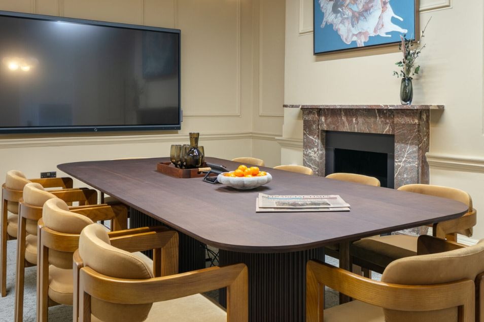 Meeting room with a dark curved table, tan wooden chairs, a marble fireplace, and a large wall-mounted screen