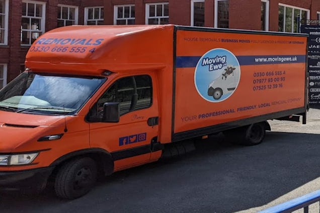 An orange Moving Ewe Ltd removal van parked on a street in front of a brick building.