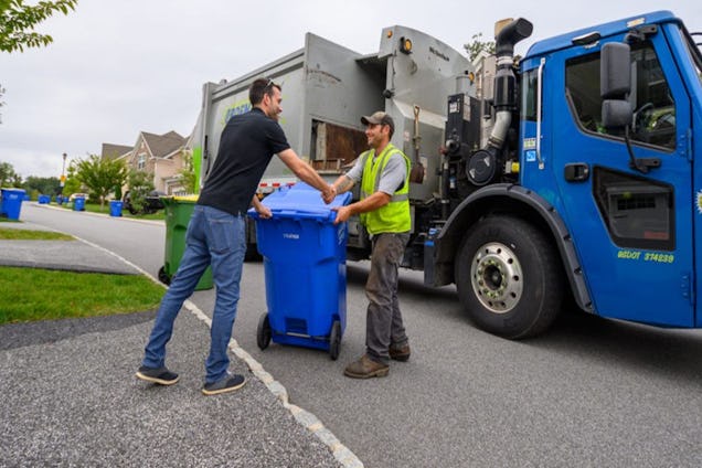A man in a black shirt hands a blue recycling bin to a worker in a yellow vest next to a blue rubbish truck.