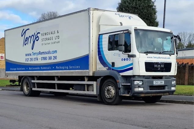 A white and blue Terry’s Removals & Storage truck parked on a street next to a brick wall.