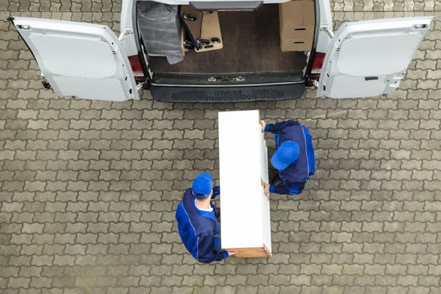 An overhead view of two workers in blue uniforms carrying a white box into the back of a van.