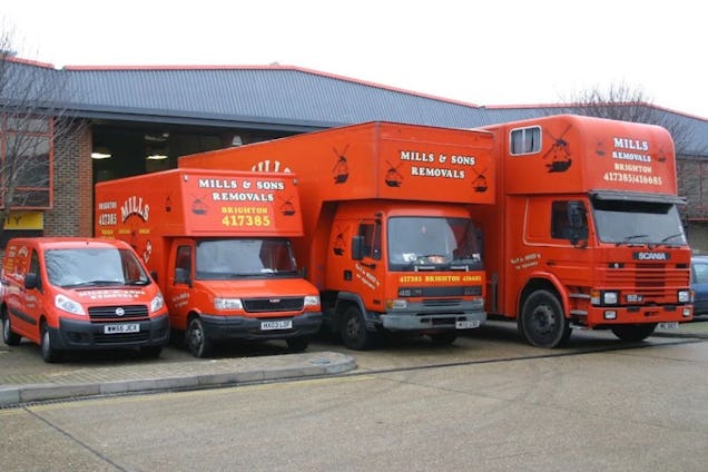 A fleet of four orange Mills Removals vans and trucks parked in a lot in front of a warehouse.