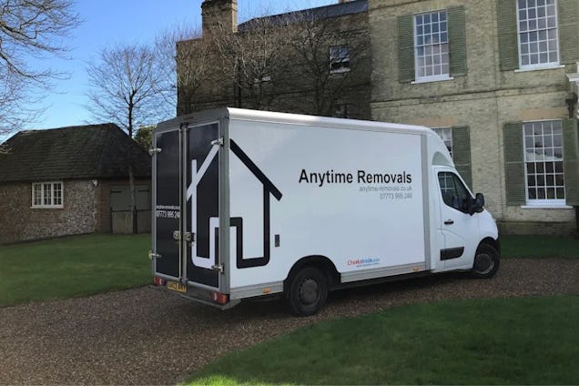A white Anytime Removals truck parked in front of a stone house with a thatched roof cottage nearby.
