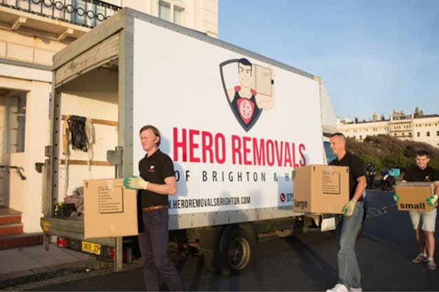 Three men carrying cardboard boxes from a Hero Removals truck parked in front of a house.