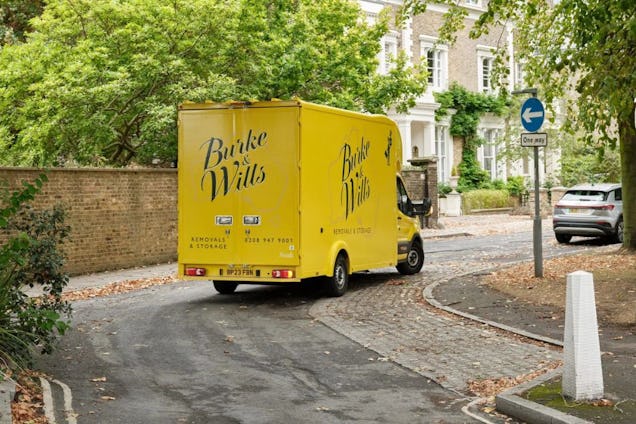 A yellow Burke & Wills removal van driving on a residential street near a stone wall and trees.