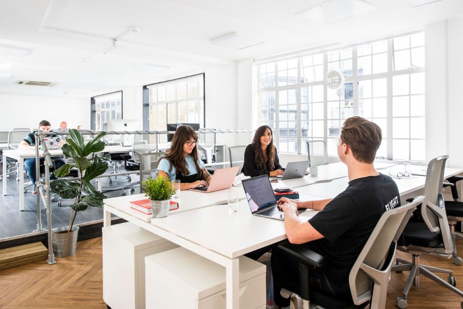 People sitting at white desks working on laptops in an office with wood floors and windows.