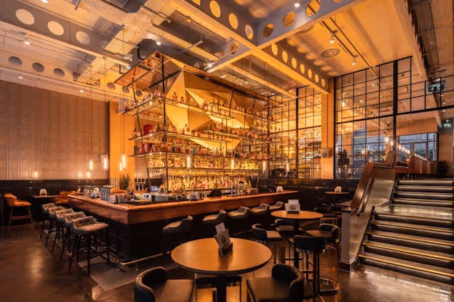 A bar interior with a wooden counter, stools, and a metal ceiling structure near a staircase.