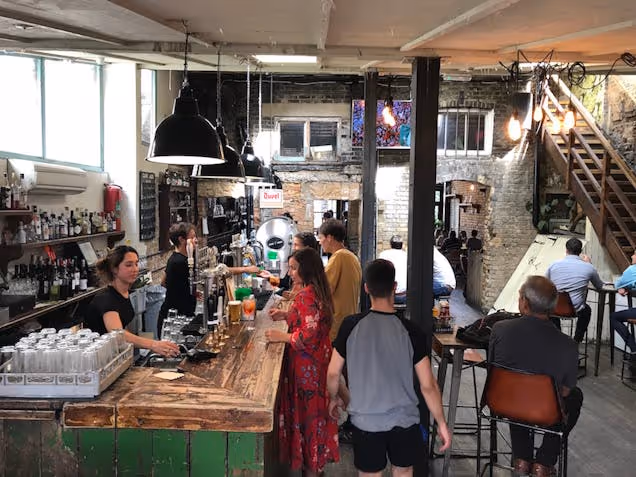 People standing and sitting at a bar counter inside a room with brick walls and a wooden staircase.