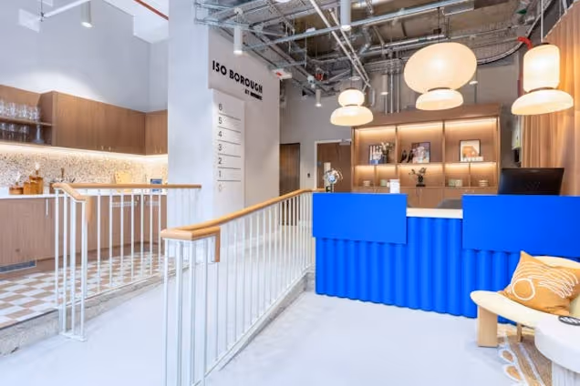 A blue reception desk next to a white staircase and seating area in an office reception area under industrial lights.