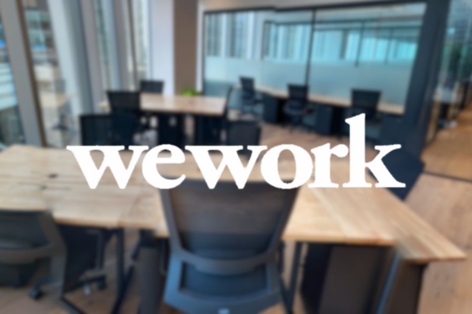 Blurry wooden office desks and black chairs in a room with glass partitions and a white "wework" logo overlay.