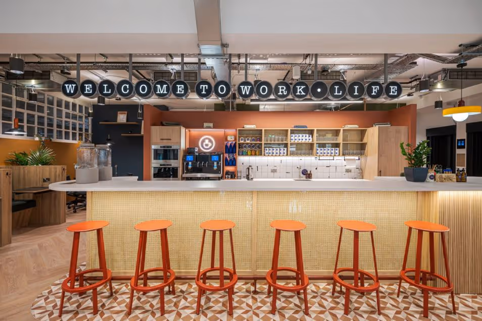A row of red stools at a wood-topped bar in an office kitchen area with tiled floors.
