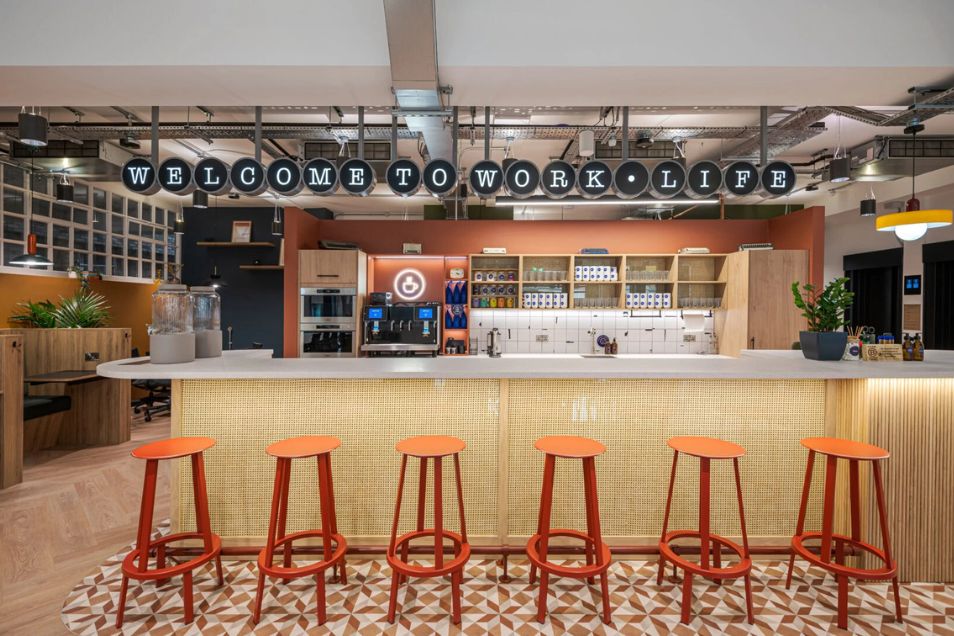 A row of red stools at a wood-topped bar in an office kitchen area with tiled floors.