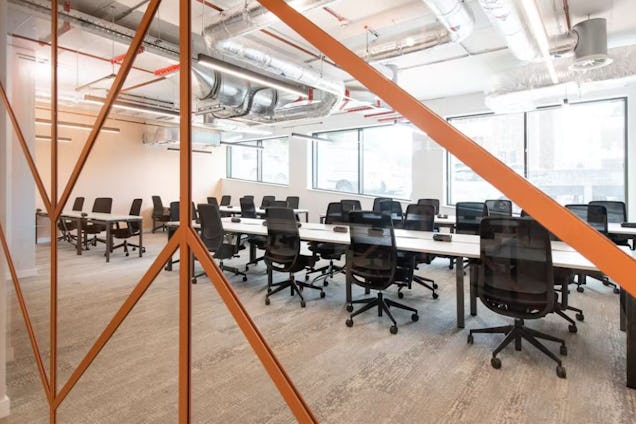 Rows of white desks with black chairs in an office featuring orange metal beams and exposed ceiling pipes.