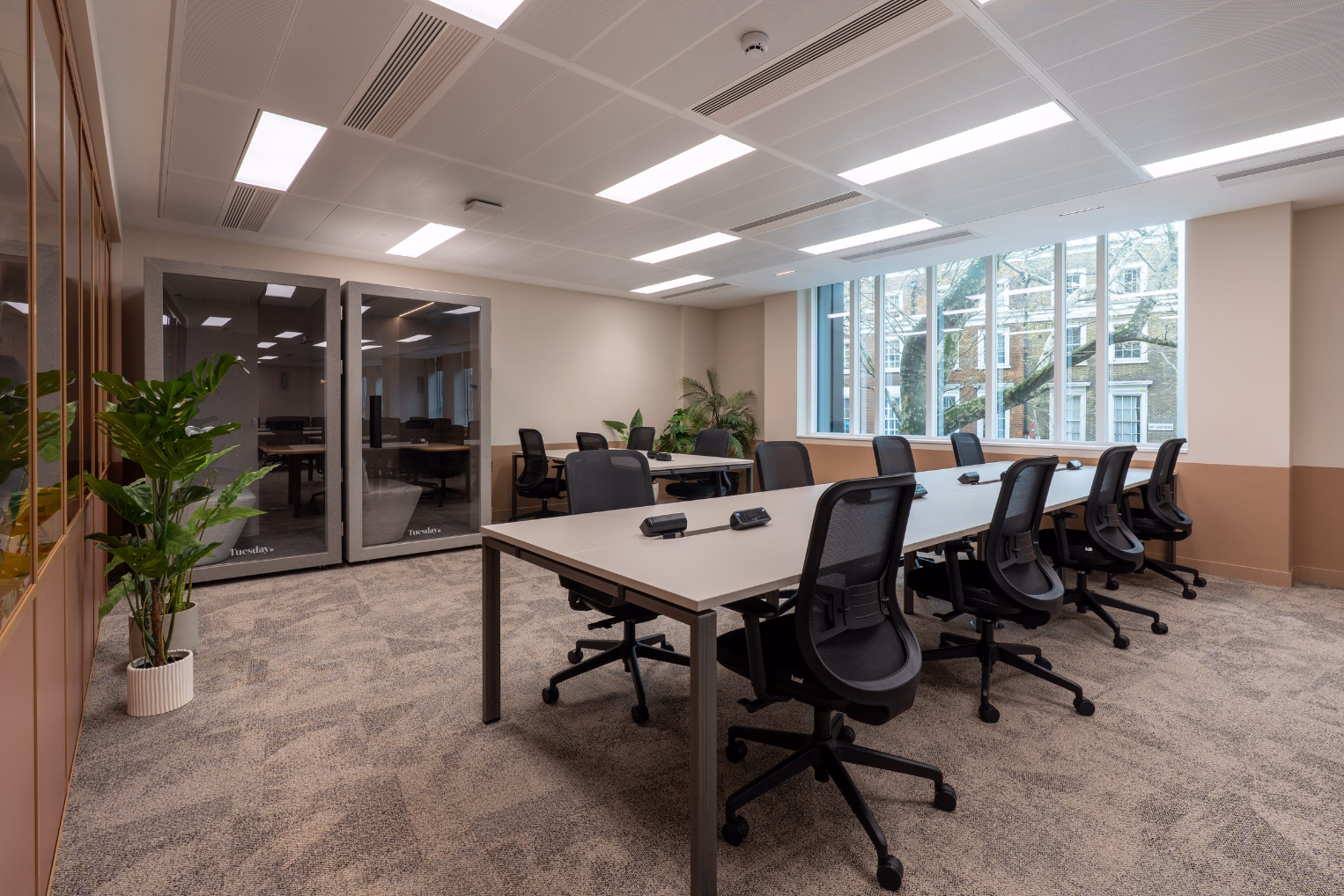 An office contains a long table with black chairs, phone booths, and a window overlooking a street.