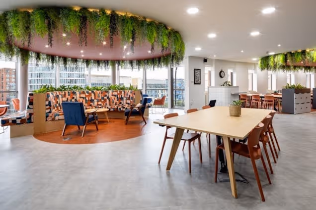 A wooden table with chairs in an office breakout area featuring hanging plants and circular seating.