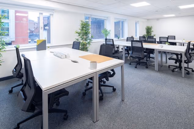 Rows of white desks with black chairs in an office with carpeted floors and windows.