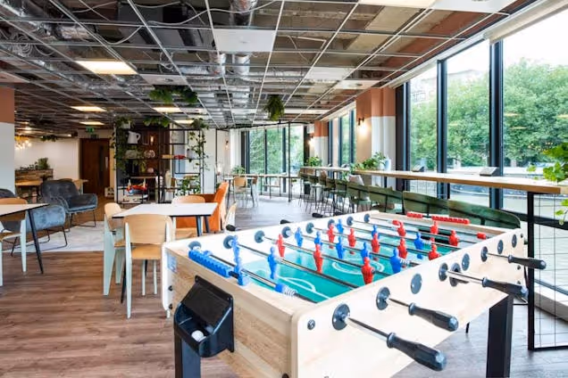 A foosball table sits in a coworking space with chairs, tables, and windows overlooking trees.