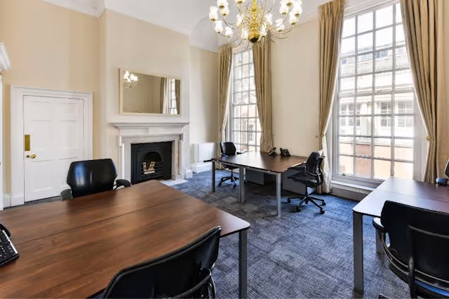 Wooden desks and black chairs in an office with a fireplace, chandelier, and windows with curtains.