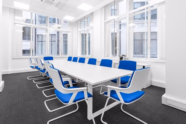 Blue and white chairs around a long white table in a meeting room with tall windows.