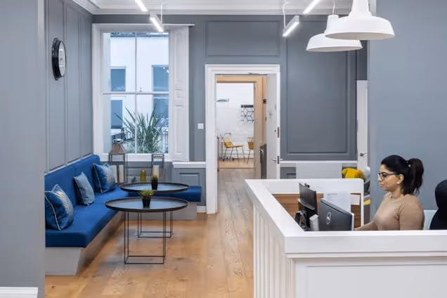 A person sits at a reception desk next to a blue sofa and a table in an office reception.