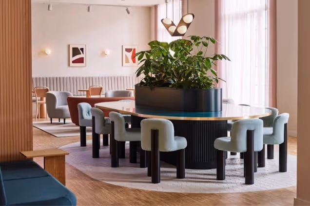 Chairs surround a circular table with a plant in the center in a lounge area with windows.