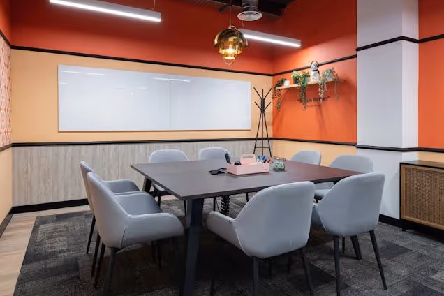 A table with grey chairs sits in a meeting room with an orange wall and a whiteboard.