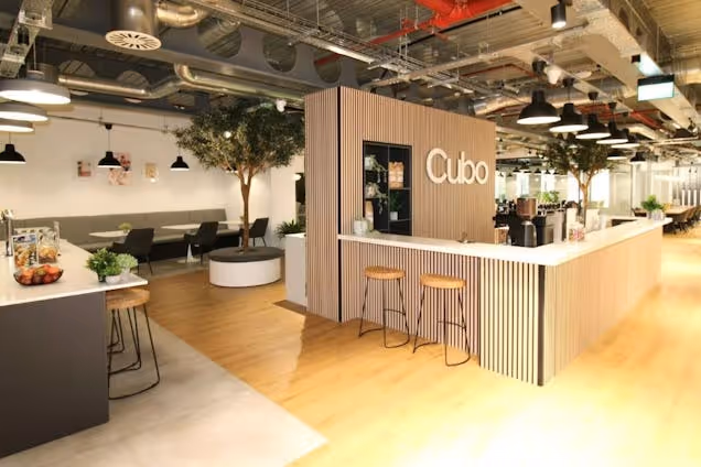 Stools at a reception counter next to a kitchen area with tables and trees in a coworking space.