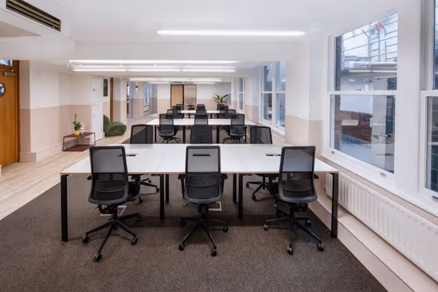 Rows of white desks with black chairs in an office with windows and carpeted floors.