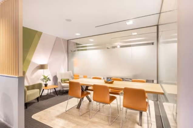 A wooden table with orange chairs in a meeting room with a rug and glass partition.