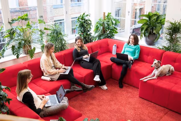 Four people on a red sofa with laptops and a dog in a breakout space with plants.