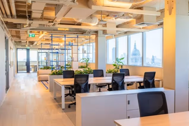 Rows of white desks with chairs in an office featuring wood floors and city views through windows.