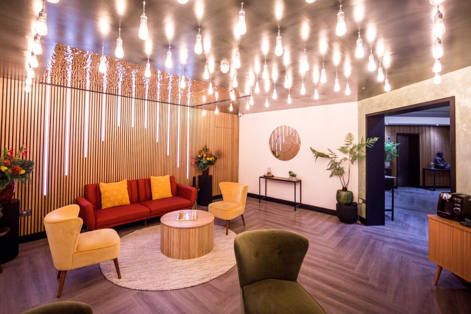 Armchairs and a sofa surround a circular table in a lobby with wood floors and lights hanging from the ceiling.