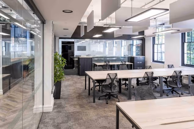 Rows of desks with black chairs in an office featuring windows and overhead lighting behind a glass partition.