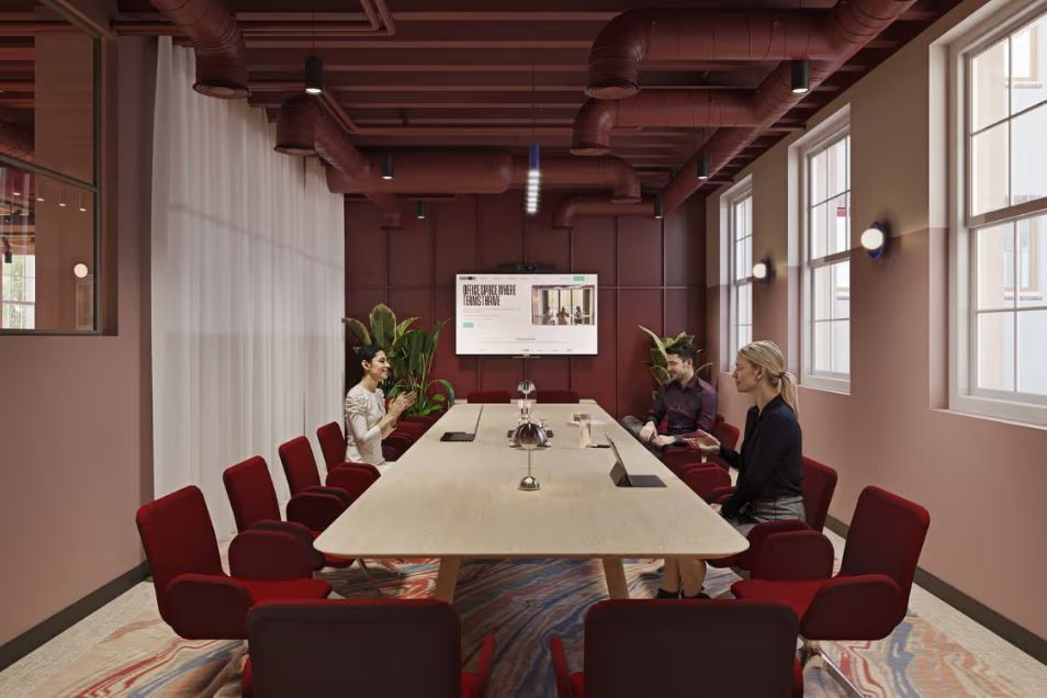 People at a long table, talking in a meeting room with red chairs and windows.