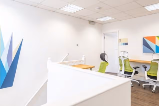 Desks and green chairs are in an office space with white walls next to a staircase.