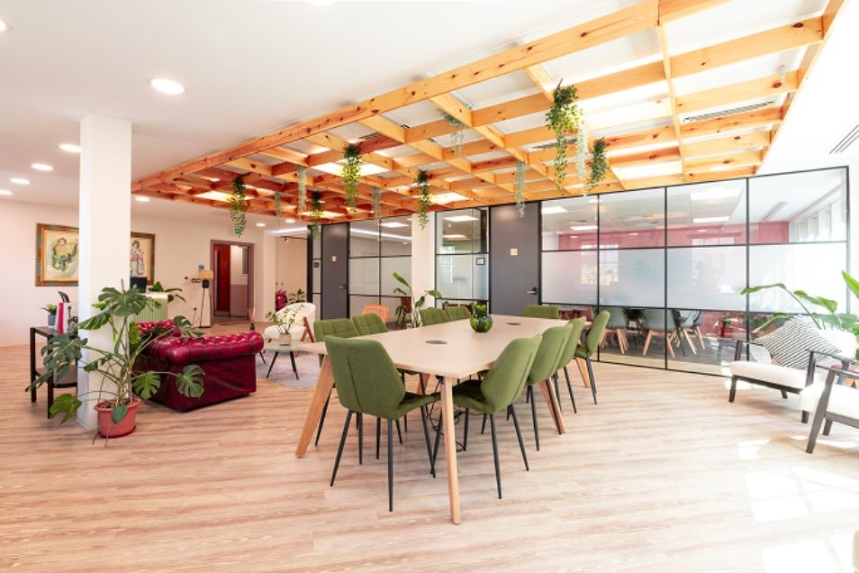 A wooden table with green chairs in a communal area with wood floors, plants, and exposed ceiling beams.