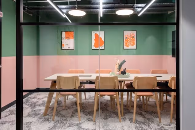 A wooden table with chairs sits in a meeting room with pink and green walls and framed art behind a glass partition.