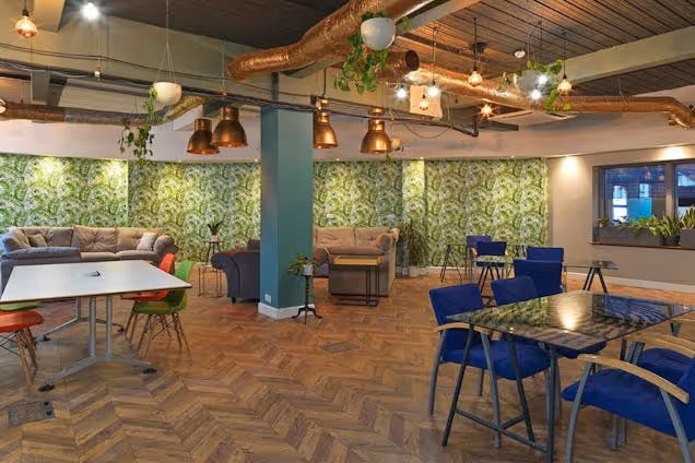 Tables and chairs sit in a coworking space featuring wood floors, plant wallpaper, and exposed ceiling pipes with hanging lights.