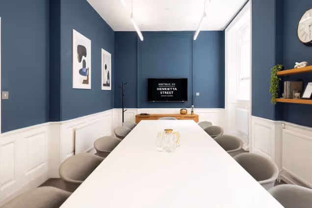 A meeting room with a white table and grey chairs in front of a blue wall and a television.