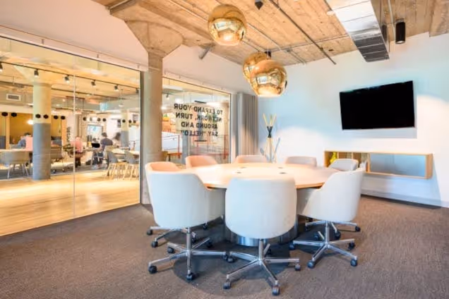 White chairs around a circular table in a meeting room with a television and glass wall.